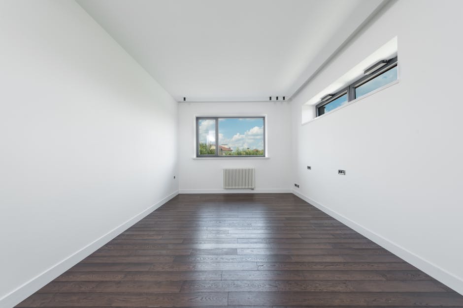 A spacious, empty room with white-painted walls and a smooth, dark wooden floor. The room features a large double-glazed window on the far wall, allowing natural light to illuminate the space and providing a view of a blue sky with scattered clouds and greenery outside. To the right, there are three narrow horizontal windows with black frames, contributing additional light and ventilation. Beneath the main window, a white radiator is mounted on the wall. The room appears to be freshly cleaned, with no visible dust or clutter, showcasing a pristine, hygienic environment suitable for domestic cleaning or surface cleaning services by Kennington Cleaners. The clean, minimalist aesthetic highlights the importance of proper maintenance and surface care in residential spaces.