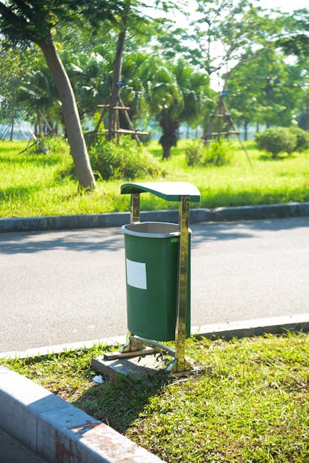 A green waste disposal bin with a white rectangular label, mounted on a metal stand with a curved top, positioned on a grassy verge beside a paved road. The bin appears clean and well-maintained. In the background, there are lush green trees and shrubbery, suggesting a park or natural setting. The scene is bright with natural sunlight, highlighting the vibrant greenery and the neat, tidy appearance of the area. This outdoor setting exemplifies environmental cleanliness and waste management, though not directly related to interior cleaning. For professional surface cleaning or deep cleaning services, visit Kennington Cleaners at kennington-cleaners.co.uk.