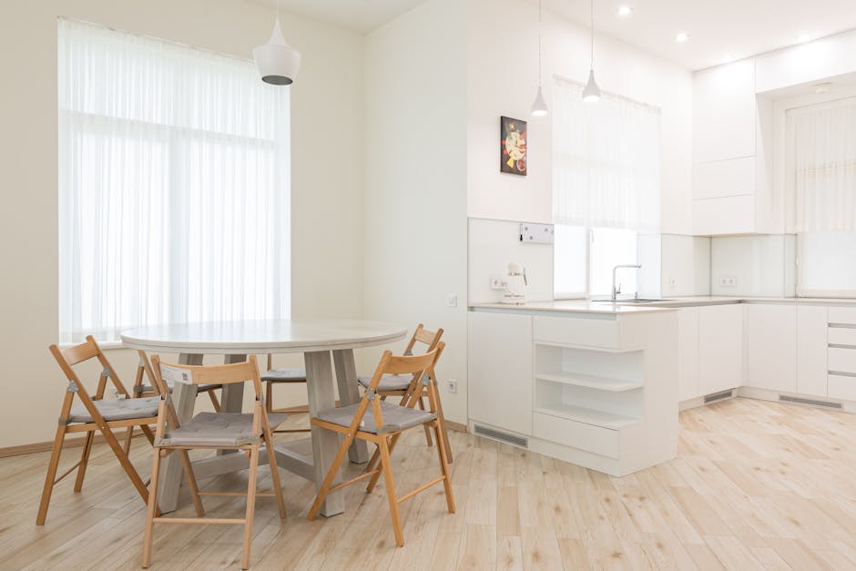A spacious, empty room with white-painted walls and a smooth, dark wooden floor. The room features a large double-glazed window on the far wall, allowing natural light to illuminate the space and providing a view of a blue sky with scattered clouds and greenery outside. To the right, there are three narrow horizontal windows with black frames, contributing additional light and ventilation. Beneath the main window, a white radiator is mounted on the wall. The room appears to be freshly cleaned, with no visible dust or clutter, showcasing a pristine, hygienic environment suitable for domestic cleaning or surface cleaning services by Kennington Cleaners. The clean, minimalist aesthetic highlights the importance of proper maintenance and surface care in residential spaces.